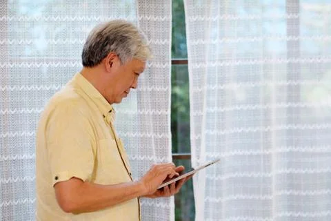 Senior asian man using  digital tablet while standing in living room at home, Stock Photos