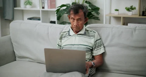 Senior Asian man using laptop while sitting on sofa in living room at home. Stockbeeldmateriaal 316018136