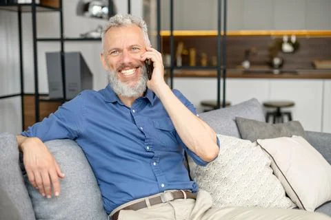 Senior bearded guy sitting down on the comfortable couch and talking on the Stock Photos