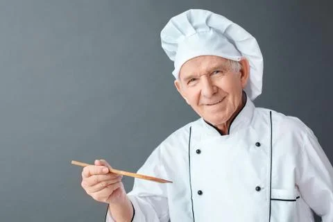 Senior chef studio standing isolated on gray holding spatula looking camera Stock Photos