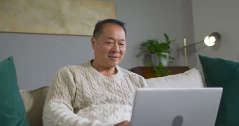 Senior Chinese man sitting on sofa wearing cream sweater, reacting to screen, Stock Footage 337730546