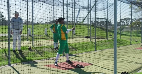 Senior coach tossing baseball at mid adult man practicing swing in batting cage, 動画素材 344253146