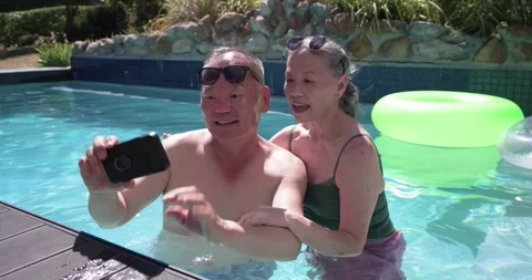 Senior couple at pool man extending phone taking selfie woman in swimsuit Stock Footage 343838585