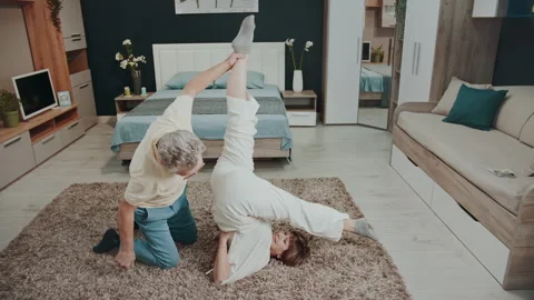 Senior couple practicing assisted yoga in a cozy bedroom. 스톡 동영상 327538523