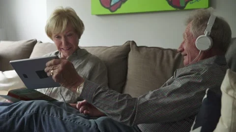 Senior couple relaxing, using digital tablet and reading book on living room sof Stock Footage 220365022