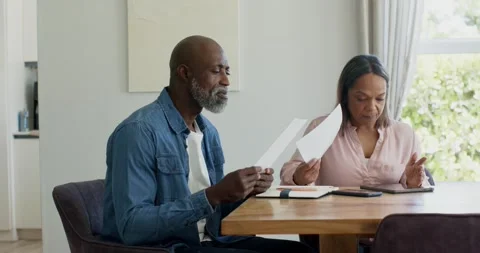 Senior couple sitting at dining table at home reviewing papers and tapping Stock Footage 338435293