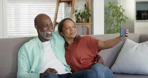 Senior couple sitting on grey couch, woman extending smartphone, man putting Stock Footage 337932985