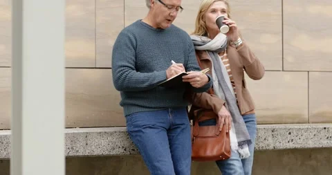 Senior couple strolling to ledge by beige building leaning scribbling notes and Stockbeeldmateriaal 340972521