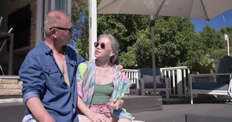 Senior couple in sunglasses sitting at backyard pool, photographer calling, Stock Footage 343367200