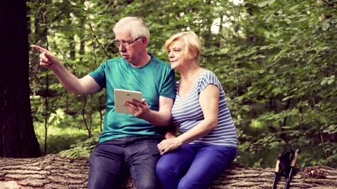 Senior couple with tablet sitting on the forest trunk and talking abut their rou Stock Footage 88427641