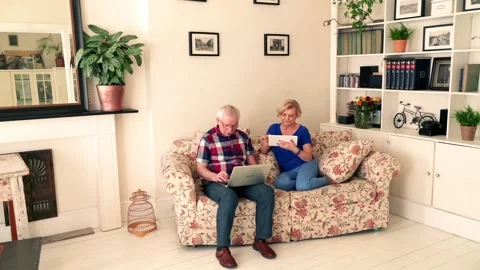 Senior couple talking and using modern laptop and tablet on sofa at home Видео 106195188