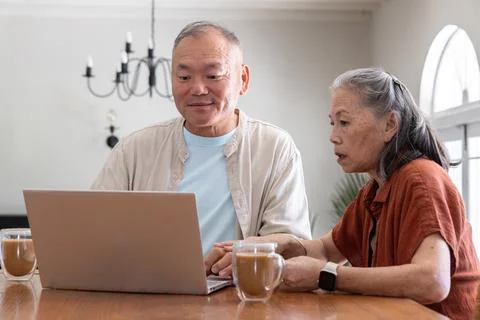 Senior couple working on laptop while sipping from clear glass mugs at wooden Fotos Stock
