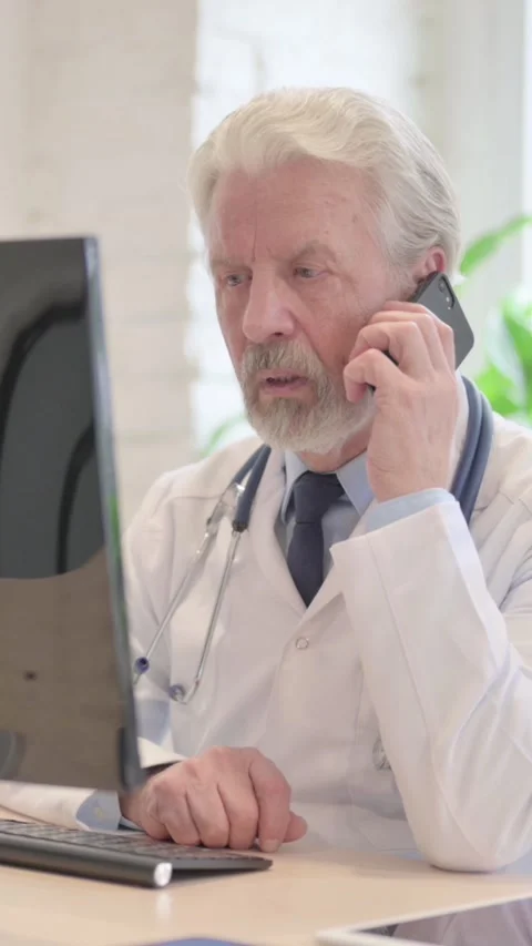 Senior Doctor Talking on Phone while Working on Computer, vertical video Vídeos de archivo 328974888