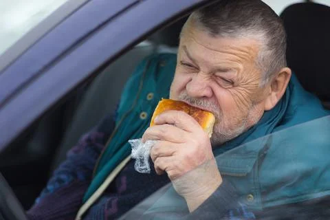 Senior driver eating patty while sitting inside his car Foto stock