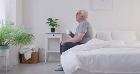 Senior elderly man sitting on bed in bedroom holding a cup of coffee or tea Stock Footage 327113092