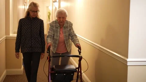 Senior elderly mother using a walker with mature daughter walking down hallway. Stock Footage 253565592