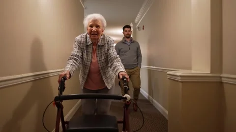 Senior elderly old mother using a walker walking down hallway with family behind Stock Footage 253404794