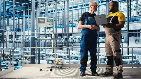 Senior engineer talks with intern in smart factory, helping trainee use software Stock Photos