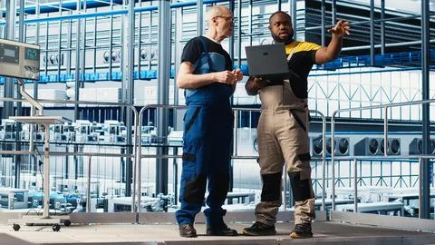 Senior engineer talks with intern in smart factory, helping trainee use software 스톡 사진
