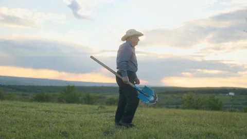 Senior Farmer Walks Through Open Field at Sunset Holding a Shovel Stock Footage 309147369
