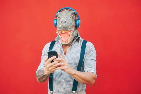 Senior fashion man wearing t-rex mask using mobile smartphone listening music Stock Photos