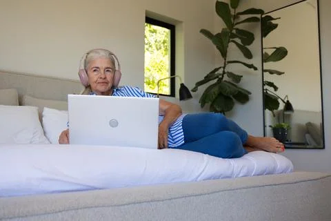 Senior female lying propped on left elbow on bed, using silver laptop and Stock Photos