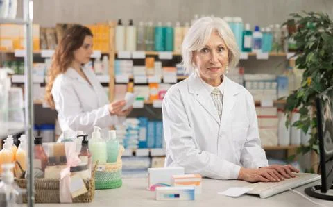 Senior female pharmacist using computer, companion at background Stockfoto's