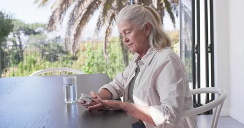 Senior female sitting at dining table near glass doors, checking white device 스톡 동영상 338356342