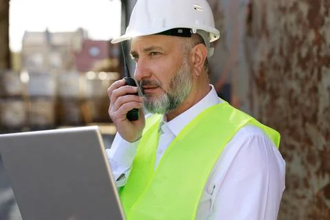 Senior foreman in protective hardhat looking at laptop and talking by walkie Stock Photos