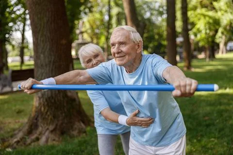 Senior gentleman does forward bend with bar rehabing injury with trainer in park Stock Photos