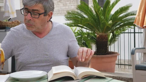 Senior gentleman sitting on the terrace eats a banana while reading a book Vídeo Stock 132871189