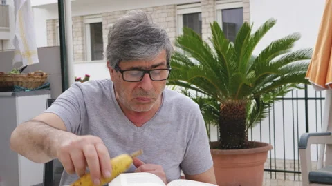 Senior gentleman sitting on the terrace eats a banana while reading a book 動画素材 132872242