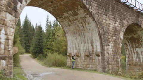 A senior gray-haired engineer stands under a very old bridge and takes Stock Footage 186747913