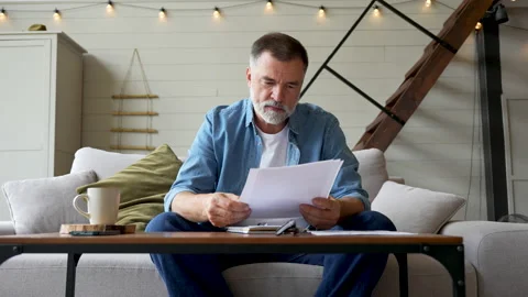Senior gray-haired man taking notes in notebook while sitting on the couch Stock Footage 222118604