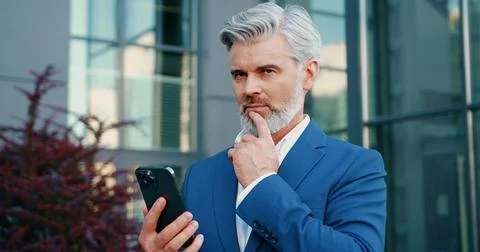 Senior grey-haired bearded businessman in a formal suit standing outdoors, using 스톡 사진