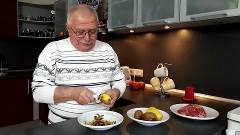 Senior grey haired man stands in modern kitchen peels potato Stock Footage 108605093