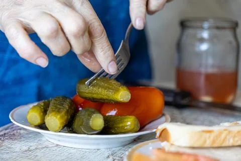 A senior hand uses a silver fork to lift a pickled cucumber from a plate with Stock Photos