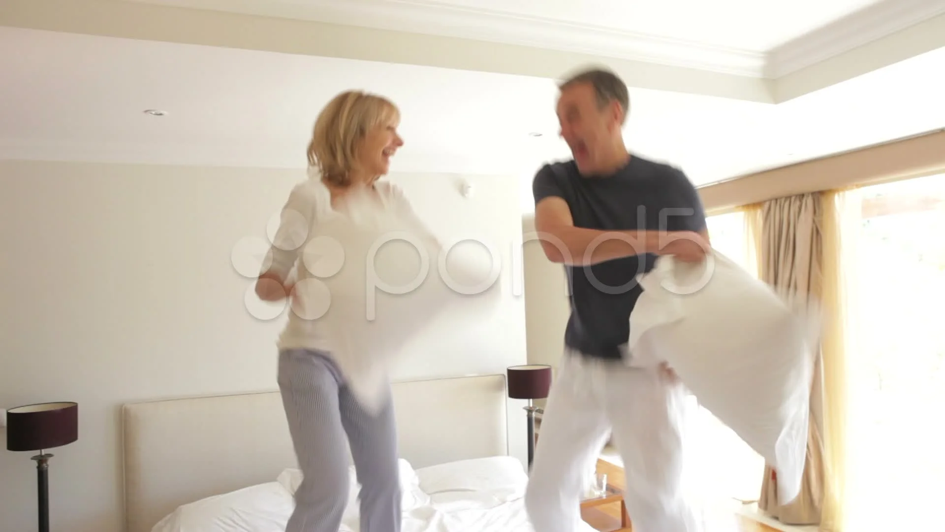 Senior husband and wife bouncing on bed having pillow fight together