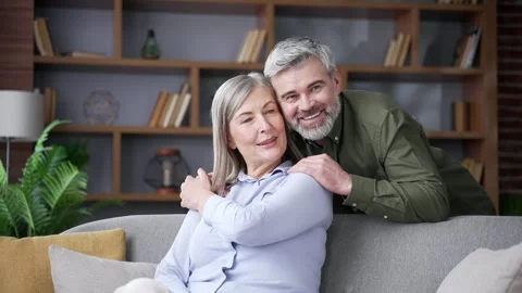 Senior husband gently hugging his wife while she sits on a sofa in the living  Stock Footage 339775901