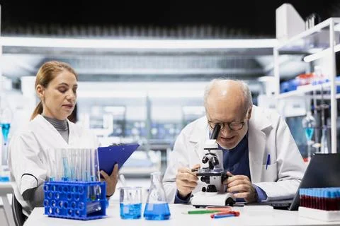 Senior lab manager using microscope next to assistant writing findings Stock Photos