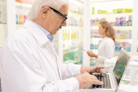 Senior male pharmacist searching for medicine in computer base by workplace Stock Photos