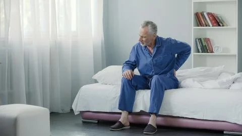 Senior male suffering sharp back pain, sick person getting up from bed, morning Stock Photos