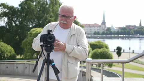 Senior man adjusts camera against river and city buildings Stock Footage 150776113
