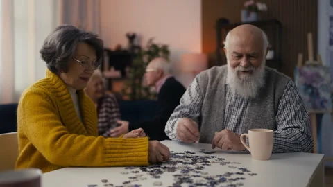Senior man and woman connect jigsaw puzzle pieces in a nursing home leisure room Stock Footage 296601853