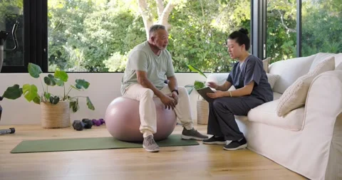 Senior man on ball leaning forward as Asian nurse holding tablet pointing Stock Footage 343242132