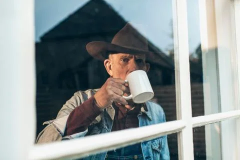 Senior man with beard and hat drinking cup of coffee behind window. Stock Photos