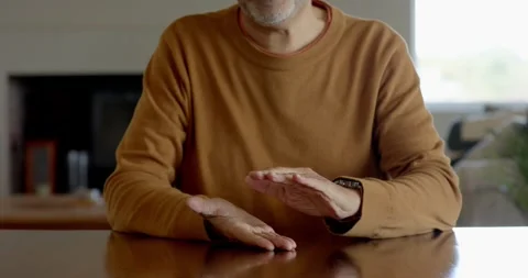 Senior man in brown sweater gesturing with hands while talking at home table Stock Footage 346028806