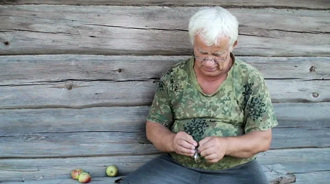 Senior man cleans a small dried fish. Wooden old house in the background, khaki Stock Footage 66202434