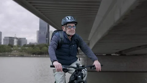 Senior Man Cycling Under Bridge Along Riverbank Wearing Helmet and Backpack Stock Footage 336044185