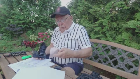 Senior man with glasses cleans camera lens at garden table Stock Footage 140966676